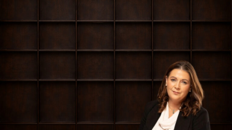 Picture of a woman wearing a black blazer and a light-colored blouse, with long brown wavy hair and pearl earrings, sat in front of a wooden panelled wall.