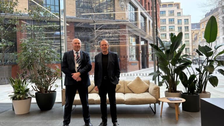 Two individuals stand in a modern lobby with large windows, potted plants, and a sofa. One wears a suit and tie, the other a dark jacket, black T-shirt, and trousers.