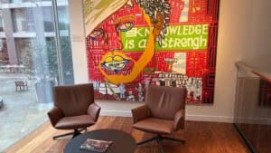 A large, colourful mural of a face with the words "Knowledge is a strength" is displayed behind two brown leather chairs and a round coffee table.