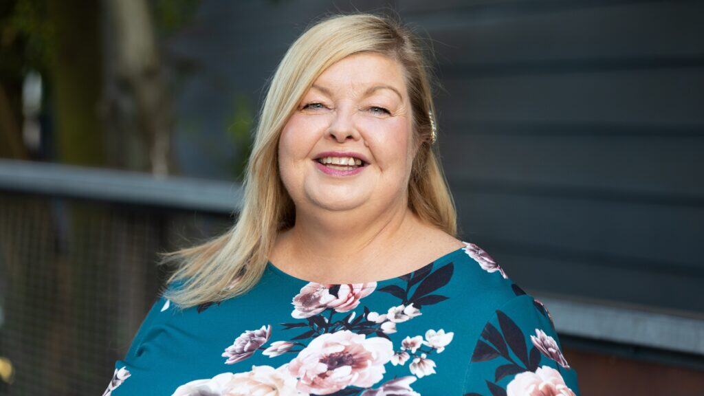 ZiaBia's Debbie Bell, smiles at the camera, wearing a teal dress with a floral pattern. She has a pearl hair clip on the side of her head. The background is blurred with a dark grey wall and some greenery.