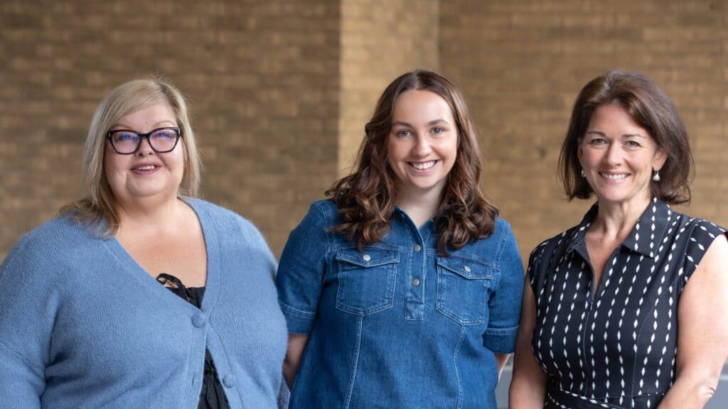Three women - ZiaBia's sustainability action standing together, smiling at the camera. The woman on the left wears a blue cardigan and glasses. The woman in the middle wears a denim dress. The woman on the right wears a black and white patterned top.