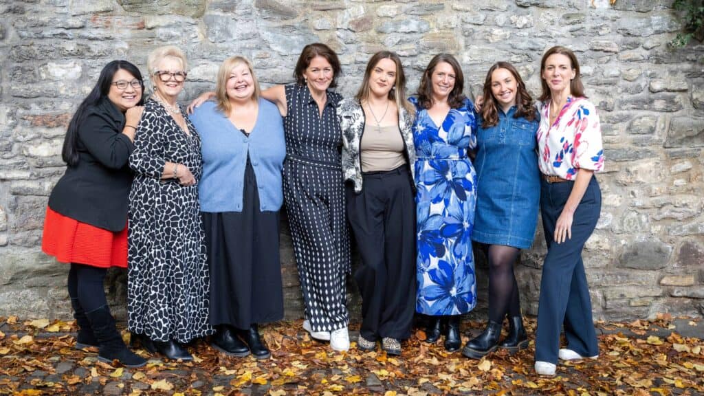 A group of eight women - ZiaBia's team - stand in front of a stone wall. They are all dressed in various outfits and are posing for the camera. The ground in front of them is covered in fallen autumn leaves.