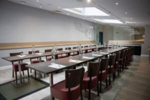 A long conference table is set with water bottles, glasses, and papers, with rows of chairs on either side in a well-lit room with skylights.