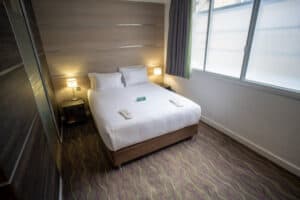 A neatly made bed with white linens and pillows sits in a hotel room with a patterned carpet and large windows with blinds.