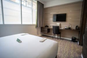 A hotel room with a bed, desk, television, and kettle. A window with blinds is on the left.