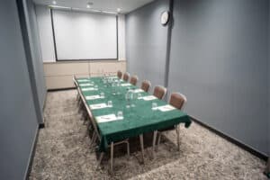 A long conference table covered with a green tablecloth, set with water bottles, glasses, and papers. Chairs are arranged around the table, and a projector screen is visible on the wall. A clock hangs on the gray wall.