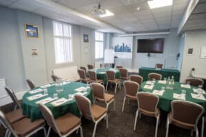 A conference room set up with round tables covered in green tablecloths, each with water bottles, glasses, and notepads. Chairs are arranged around the tables, and a projector screen with a cityscape graphic is visible on the wall.
