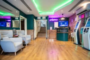 A modern reception area with teal walls, brick accents, and wood floors. It features comfortable seating, a reception desk with "THE WESLEY" logo, a "welcome" neon sign, and several self-check-in kiosks.