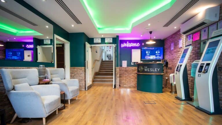 A modern reception area with teal walls, brick accents, and wood floors. It features comfortable seating, a reception desk with "THE WESLEY" logo, a "welcome" neon sign, and several self-check-in kiosks.