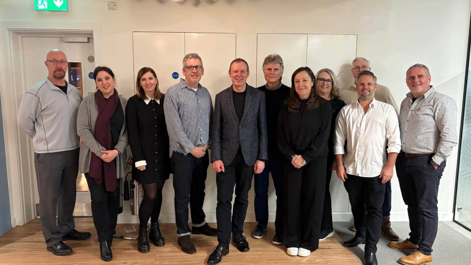 A group of eleven people stand together for a photo indoors. Most are wearing dark clothing, with some wearing grey and white tops. They are standing in two rows, with the front row slightly staggered.