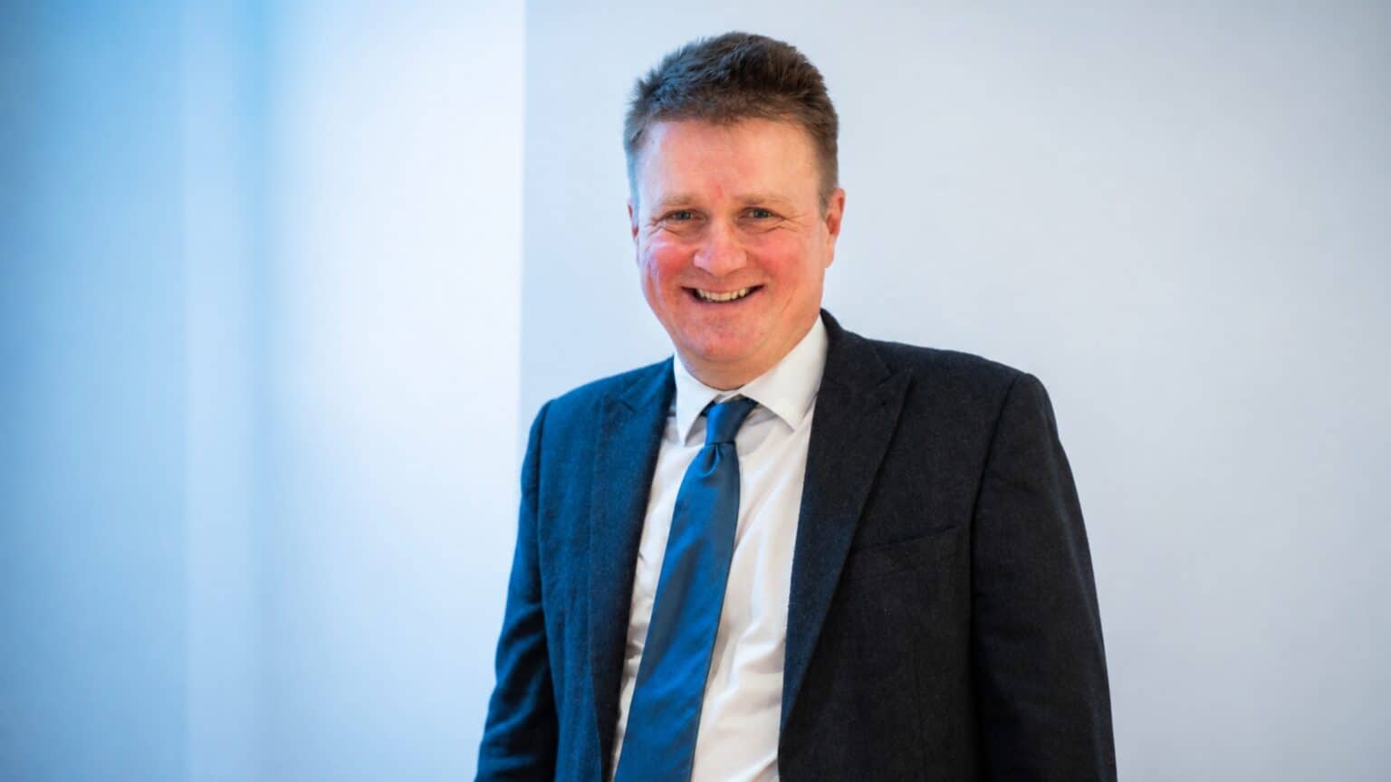 A man in a dark suit and blue tie smiles broadly at the camera against a light blue background.
